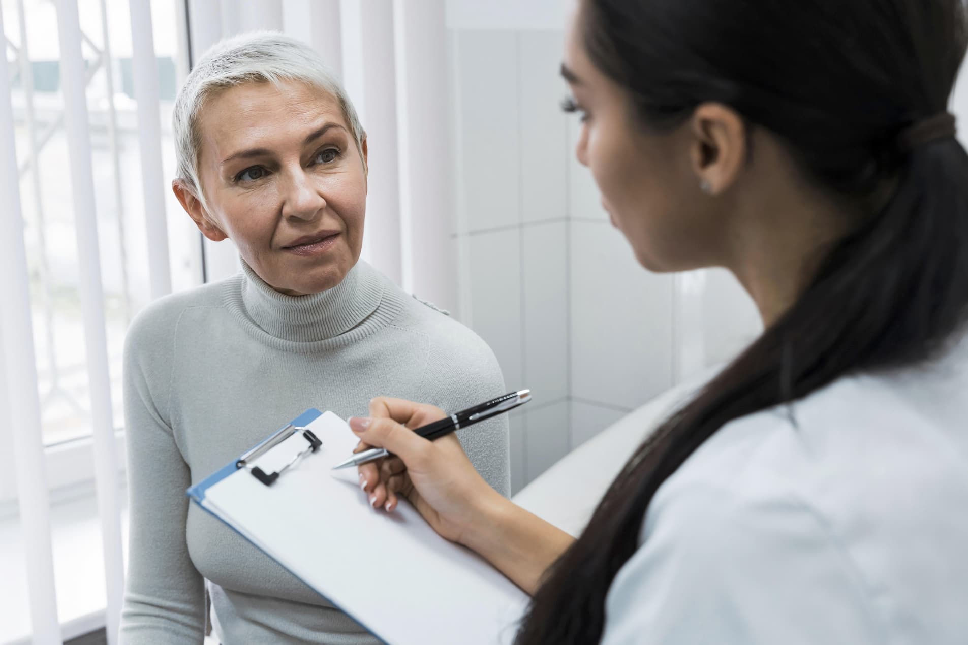 A female doctor with a clipboard talks to an elderly female patient.