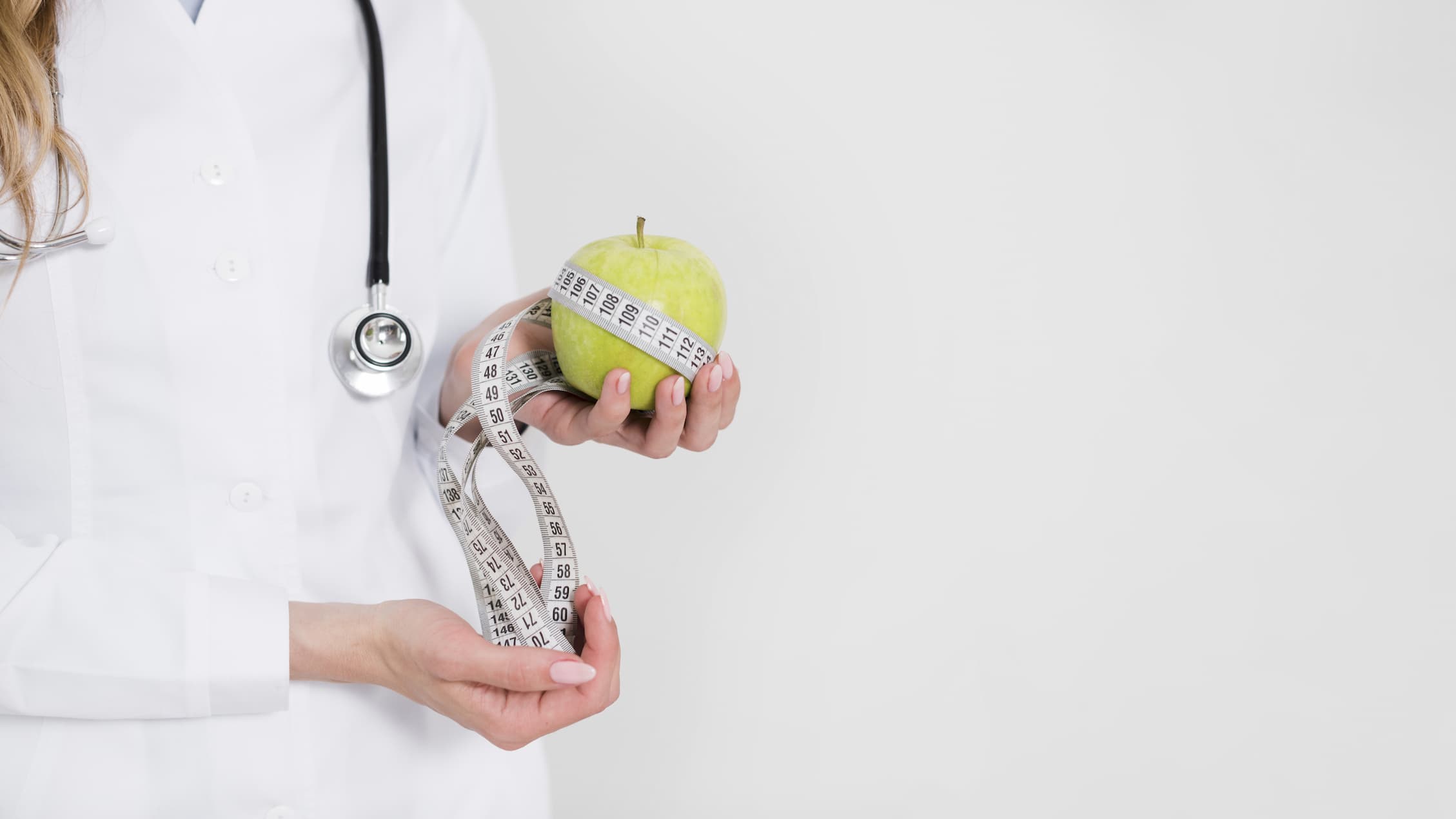A doctor in a white coat holds a green apple with a measuring tape wrapped around it.
