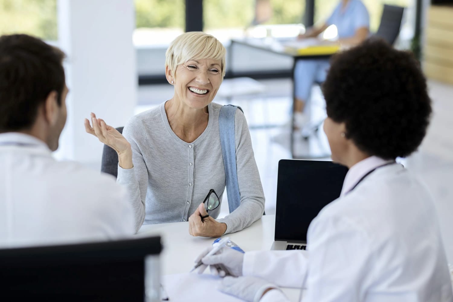A cheerful older woman with short blonde hair laughs while speaking with two doctors in a bright, modern clinic.