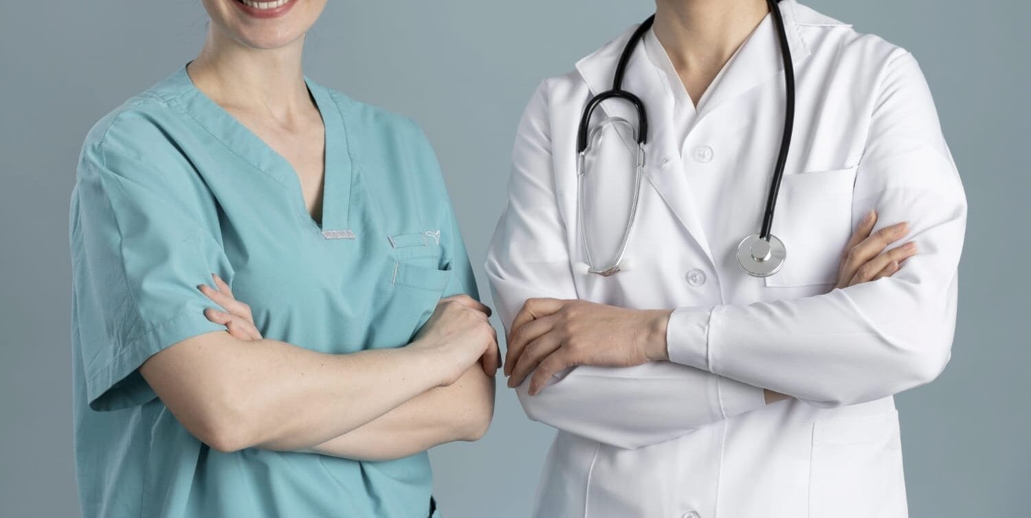 A smiling female nurse in green scrubs and an older female doctor in a white coat stand next to each other with their arms crossed.