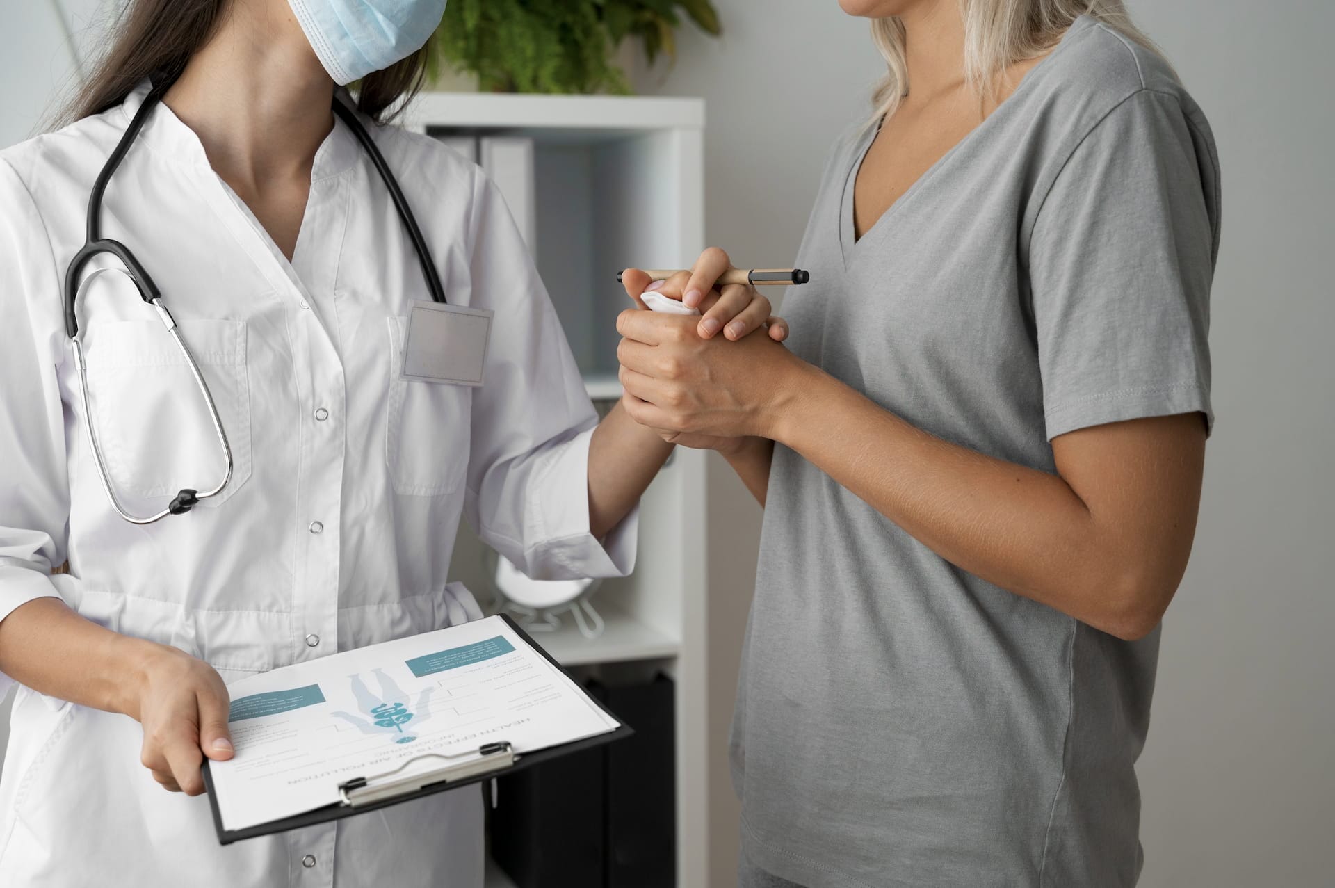 A female doctor wearing a face mask holds a clipboard and reassures a female patient by holding her hand.